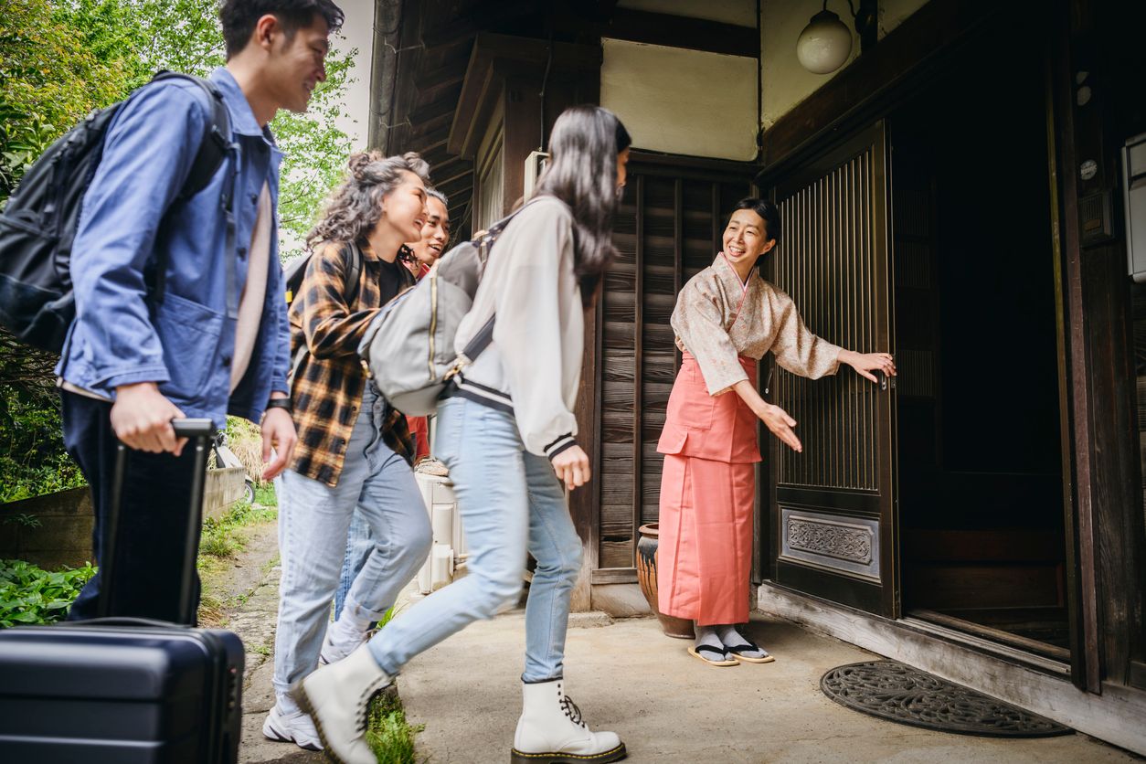 Japanese woman welcoming four backpackers with luggage, opening door, ushering them inside the building