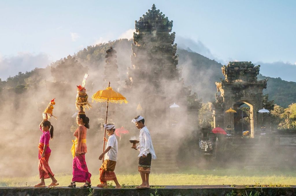 Showing traditional Balinese male and female ceremonial clothing and religious offerings as a mother and children walk to a Hindu temple (pura) in Bali
