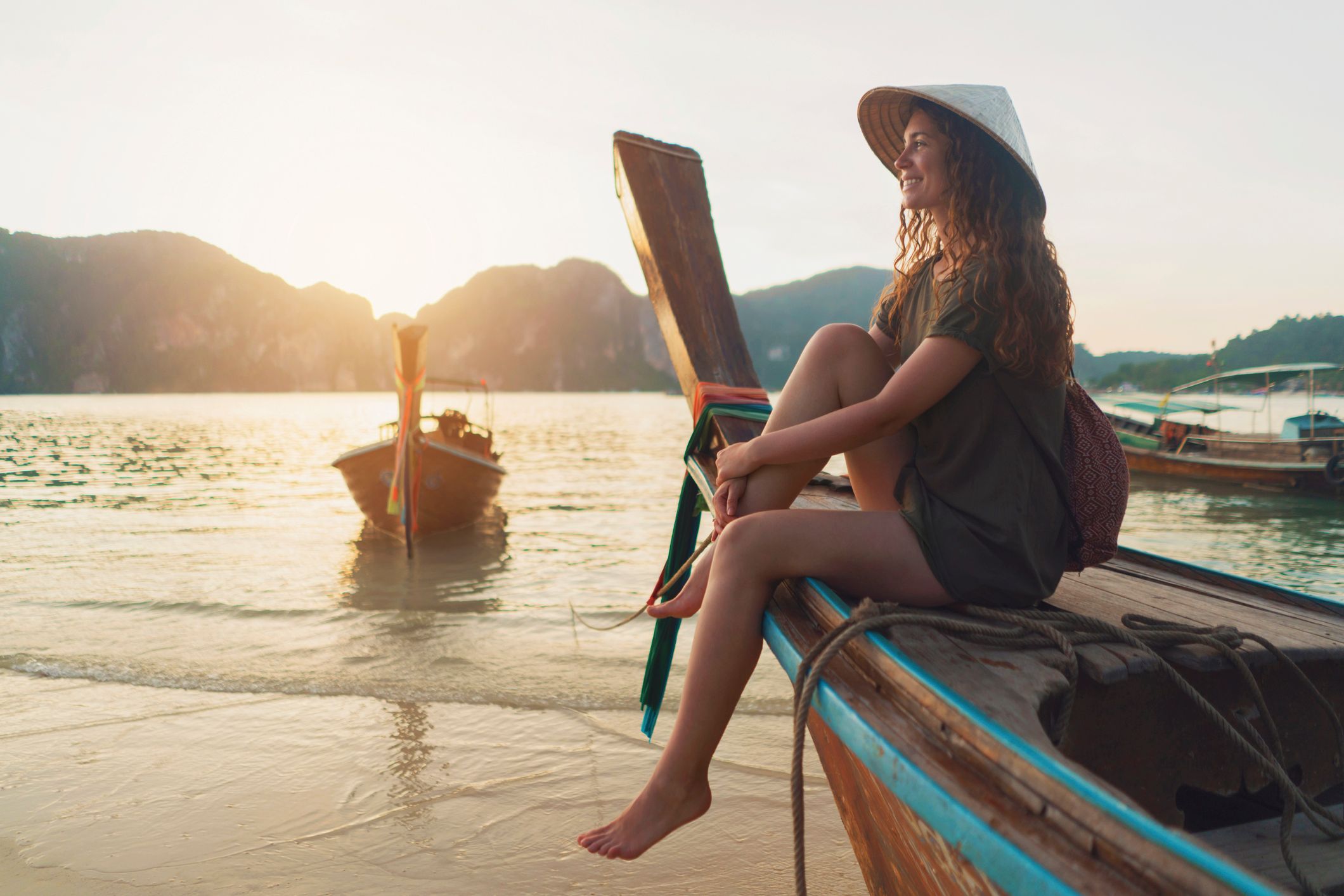 Woman with an Asian style conical hat sitting on a longtail boat and enjoying a carefree summer day on the beach.