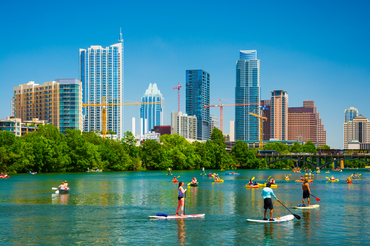 People having fun on rowboats and boards in Lady Bird Lake on a nice day with the Austin skyline in the background.