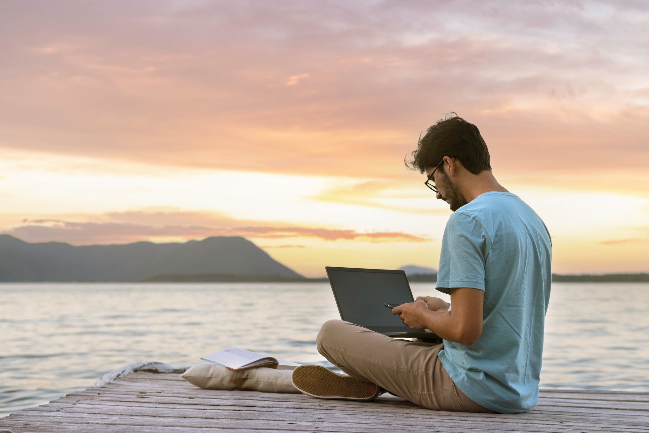 Young digital nomad man sitting on wooden pier working from a laptop